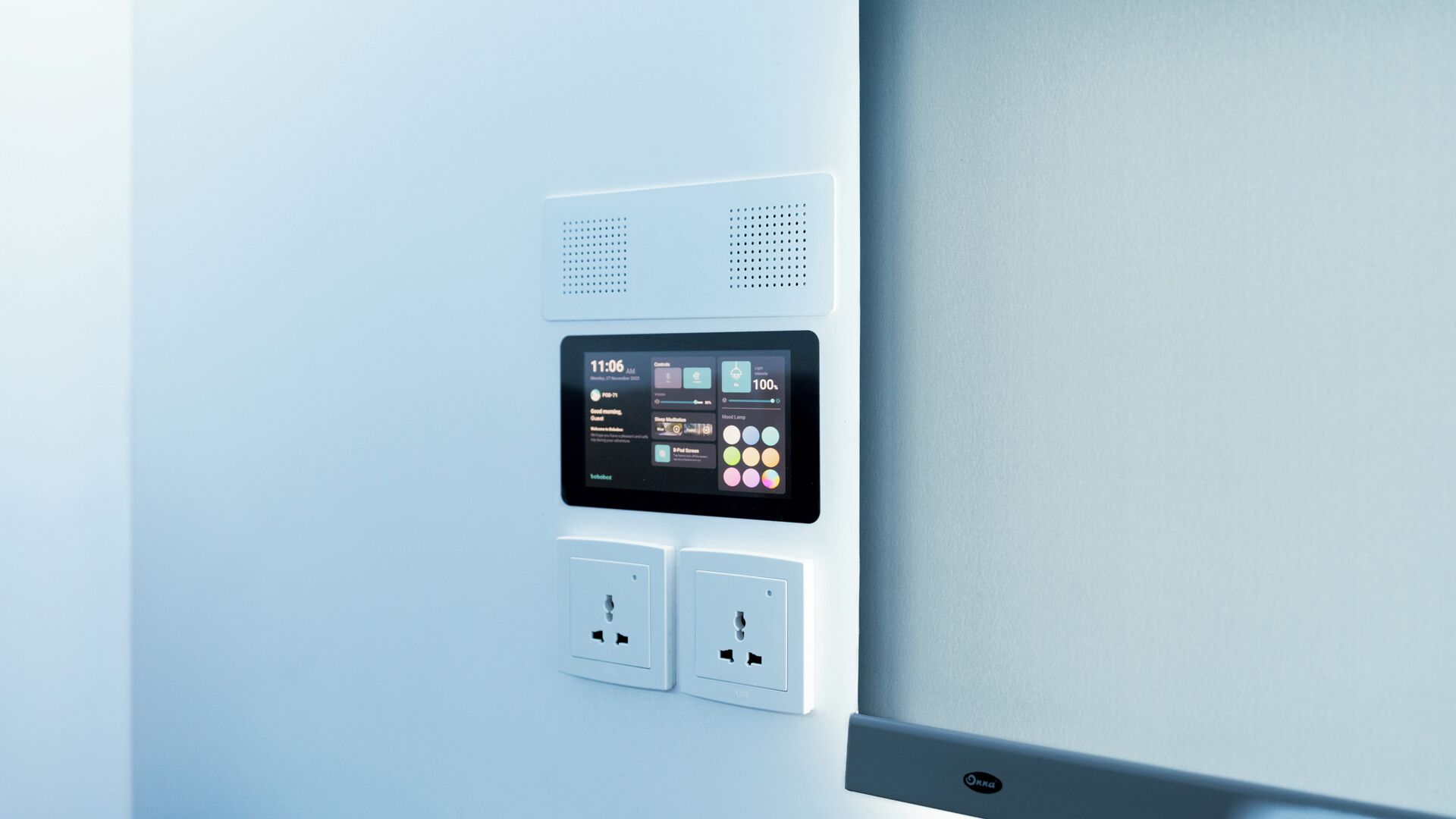 Un panel de control fijado a la pared con pantalla digital, dos enchufes y un altavoz.