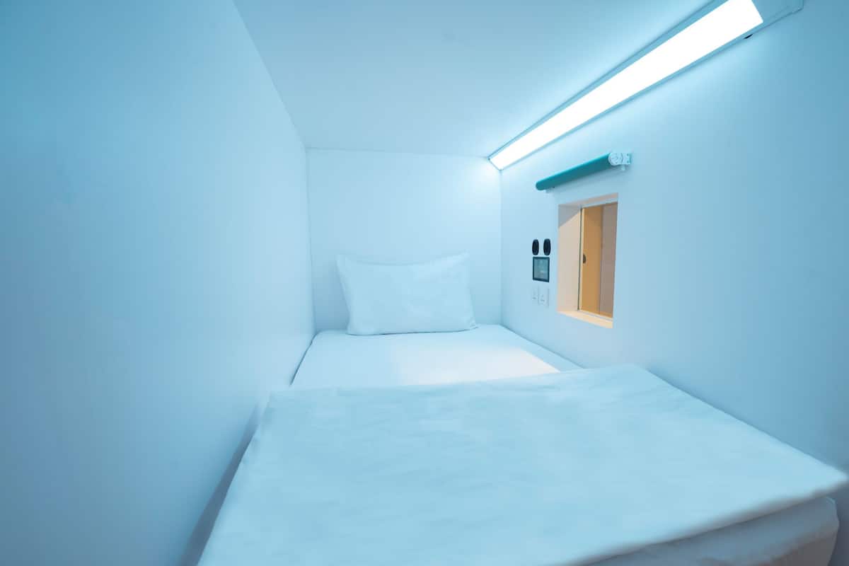 A small, narrow room with a single bed, a small window, and a wall-mounted light fixture.