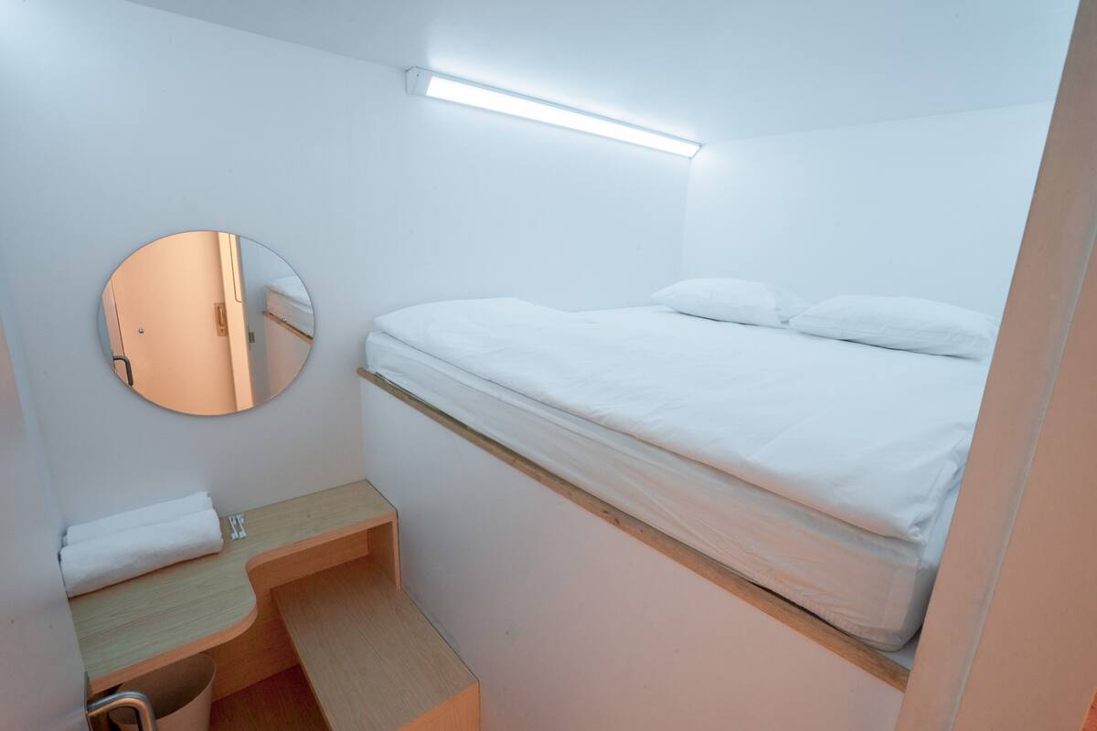 A small, clean room with a single bed, a round mirror, a wooden nightstand, and a bedside table.