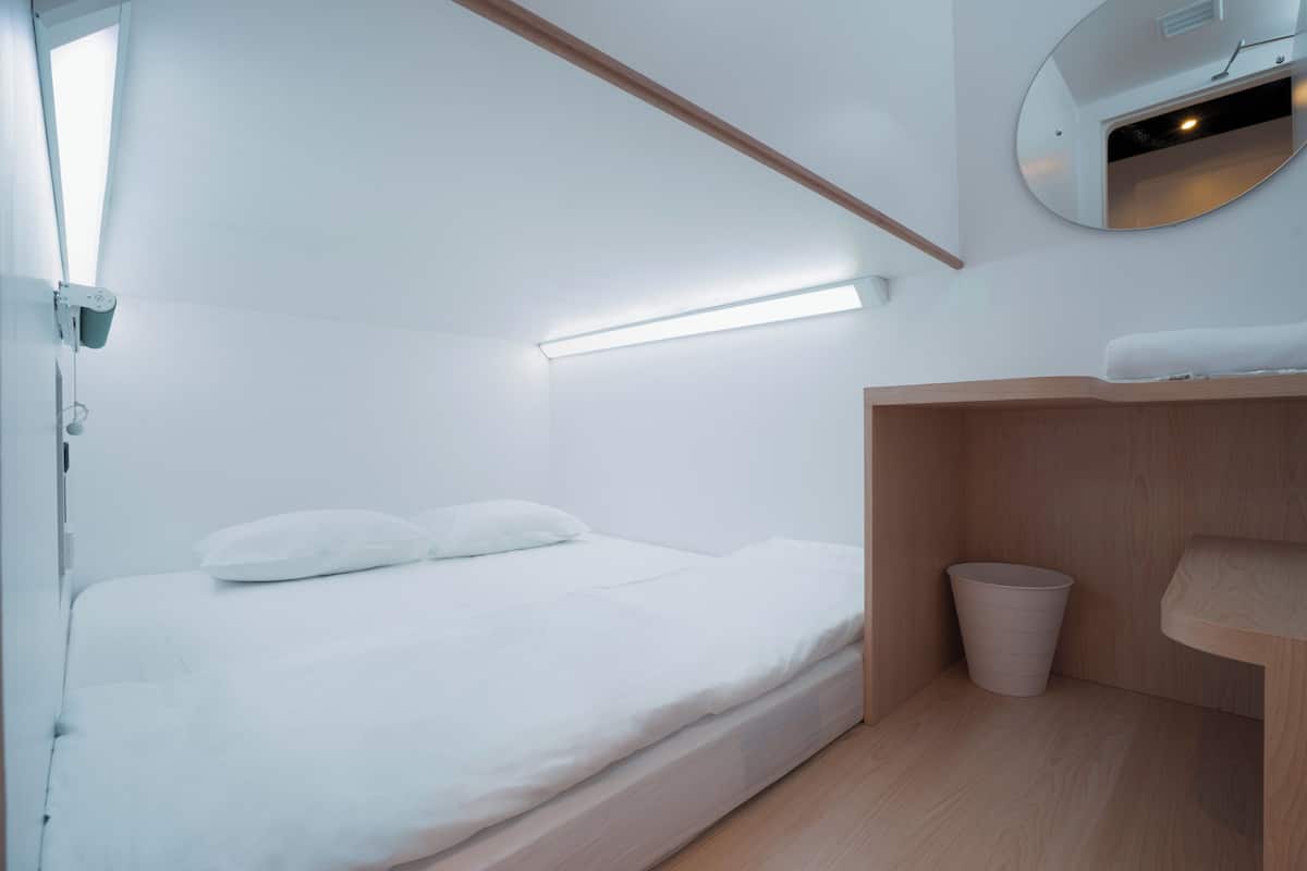 A minimalist bedroom with a bed, a bedside trash bin, and a small wooden shelf.