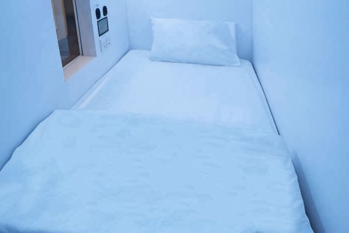 A small, single-bed room with a white bedspread and pillow, a small window with a view, and a wall-mounted control panel.