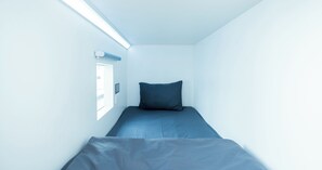 Una habitación pequeña y estrecha con una cama individual, una ventana y un panel de control en la pared.