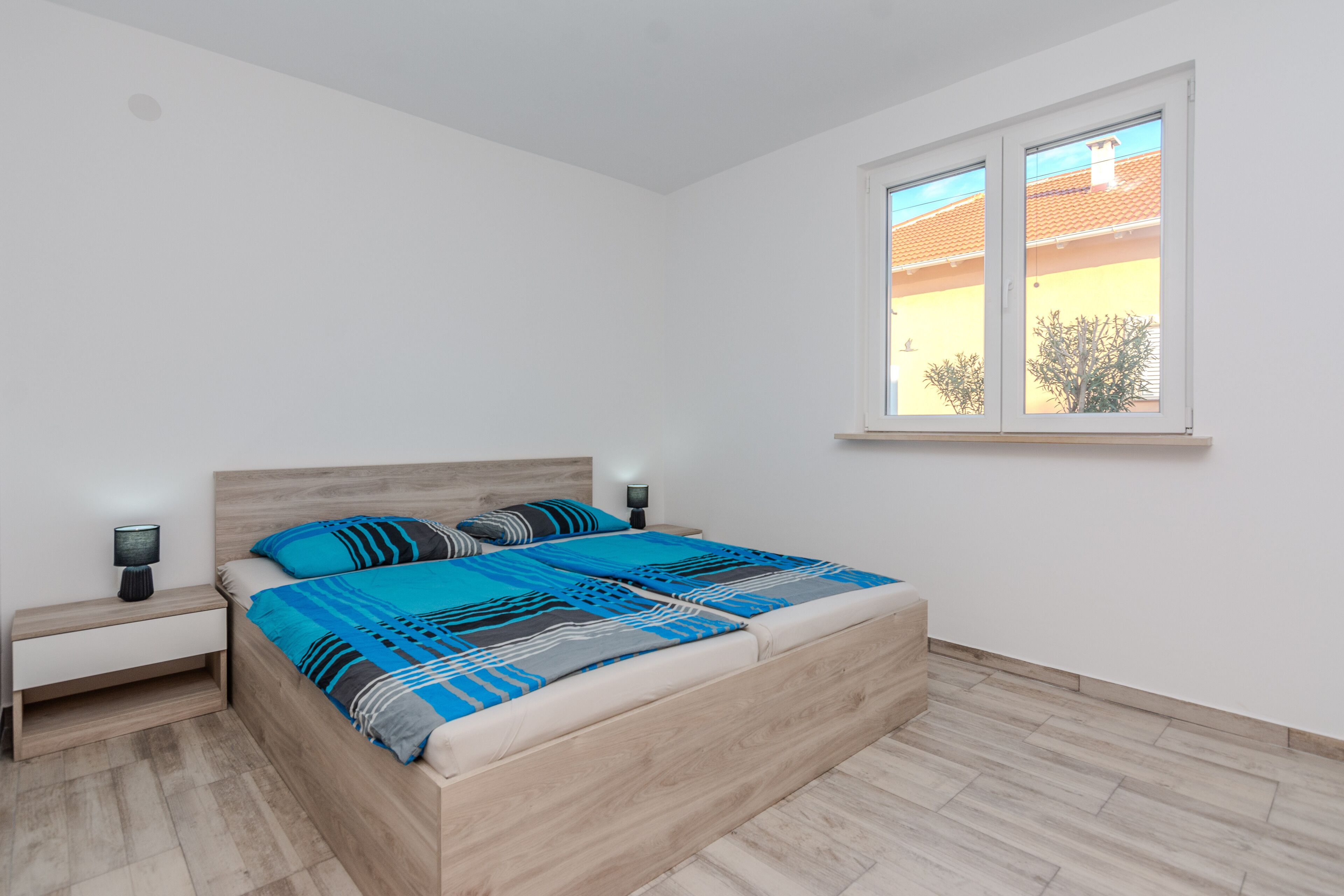 Un dormitorio con una cama de madera, una mesita de noche, una lámpara de noche y una ventana con vistas a un edificio y árboles.