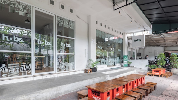 An outdoor seating area with wooden tables and benches, a Coca-Cola branded cooler, and a storefront with large windows and signage reading "h.b.s." - HUBS Hostel Yogyakarta (Yogyakarta)