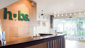 A reception desk with a wooden panel and a large green 'hbs' sign. There are clocks on the wall and a view of the outside through a glass door. - HUBS Hostel Yogyakarta (Yogyakarta)