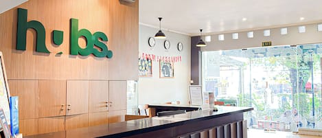 A reception desk with a wooden panel and a large green 'hbs' sign. There are clocks on the wall and a view of the outside through a glass door.