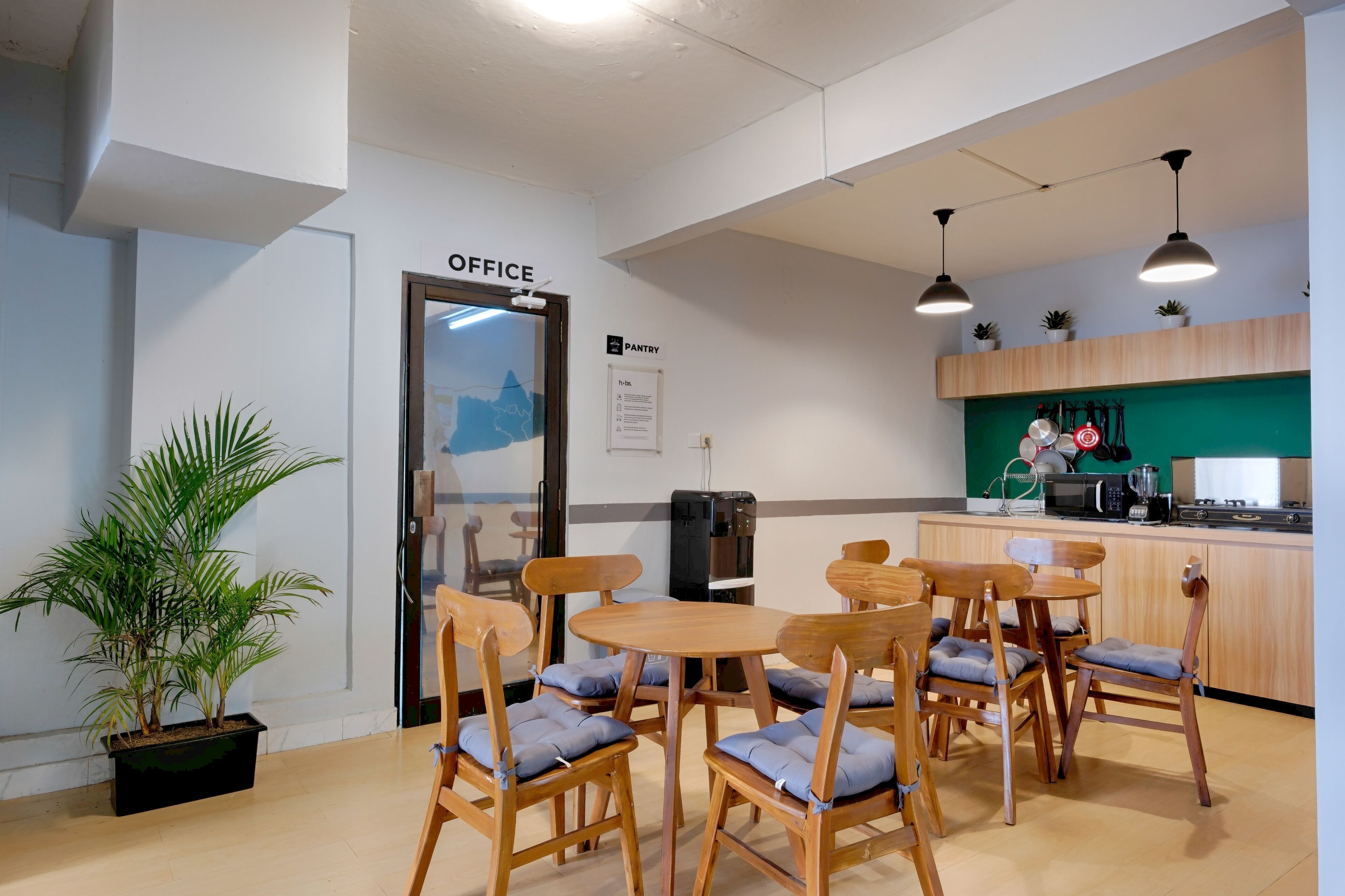 Un comedor con sillas de madera y una mesa, una kitchenette con salpicadero verde, y un letrero que indica la ubicación de la oficina.