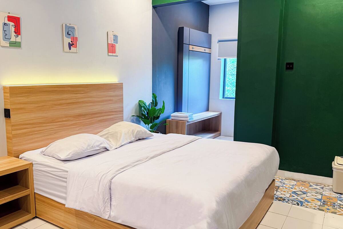 A modern hotel room with a wooden headboard, a bed with white linens, a bedside table, a green wall, and a window.