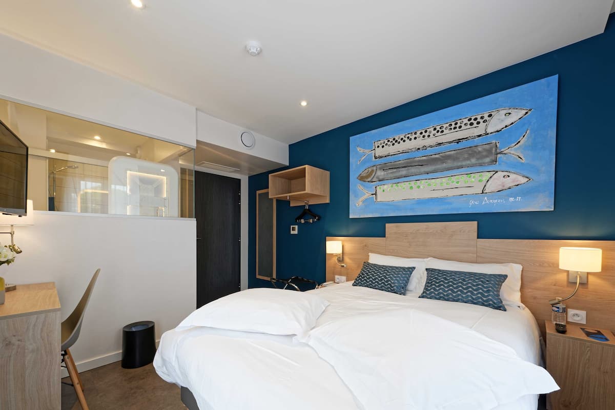 A modern hotel room with a large bed, two bedside tables with lamps, a wall-mounted artwork of fish, a desk with a chair, and a view of the bathroom.