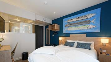 A modern hotel room with a large bed, two bedside tables with lamps, a wall-mounted artwork of fish, a desk with a chair, and a view of the bathroom.