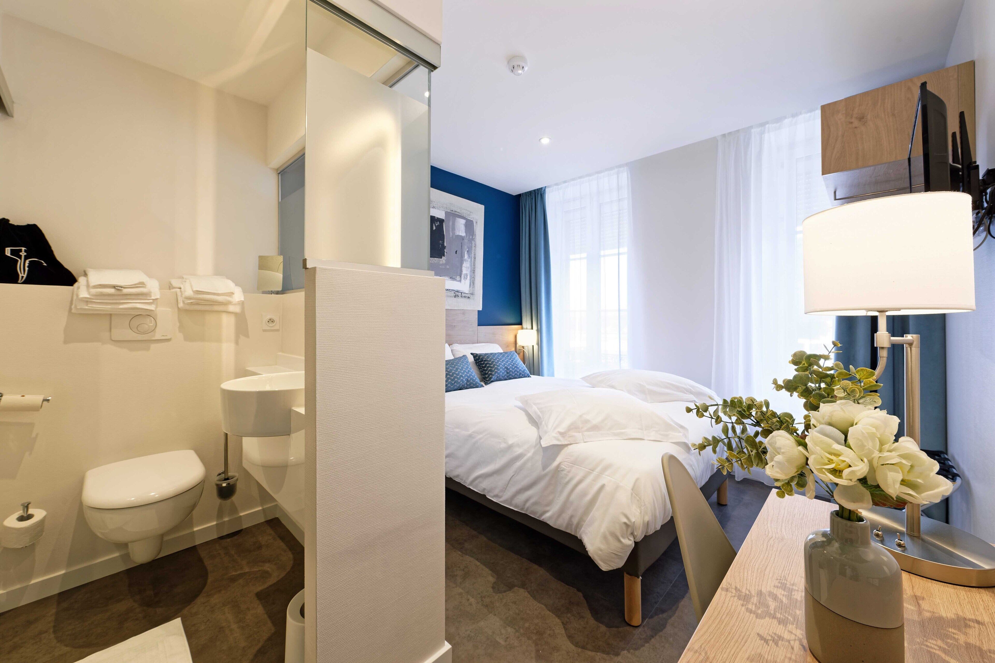 A hotel room with a bed, a bathroom, a desk with a vase of flowers, and a window with curtains.
