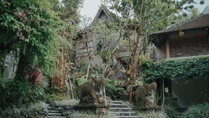 Une maison traditionnelle avec un escalier en pierre menant à une porte en bois, entourée d’une végétation luxuriante et de statues en pierre.