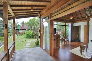 Un porche de madera cubierto con suelo de cuadros, una puerta de vidrio que da a una habitación con escritorio y silla, y vista a un jardín con una estructura de techo de paja.