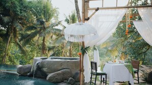 Un espace repas aménagé au bord de la piscine, avec une table dressée pour deux personnes, entouré de rochers et de verdure.