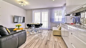 Een moderne woonkamer met een bank, een eettafel met stoelen, een keuken met een kookplaat, en een aan de muur bevestigde televisie.