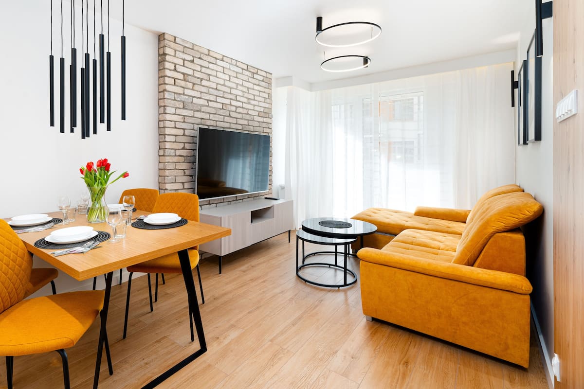 Een moderne woonkamer met een houten eettafel, oranje stoelen, een bank en een televisie bevestigd aan een bakstenen muur.