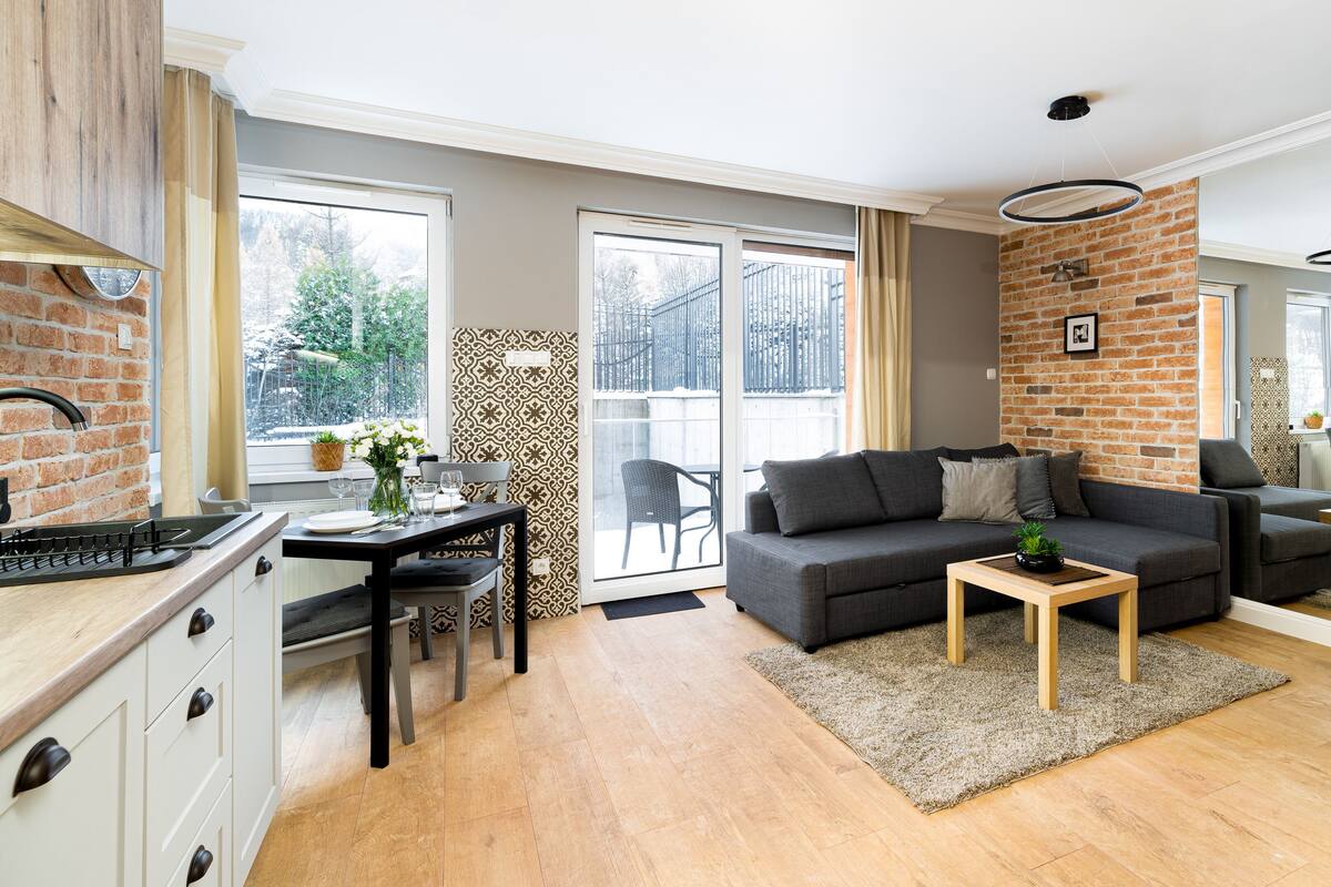 Een moderne woonkamer met een grijze bank, een klein houten bijzettafeltje en een eethoek met een tafel en stoelen.