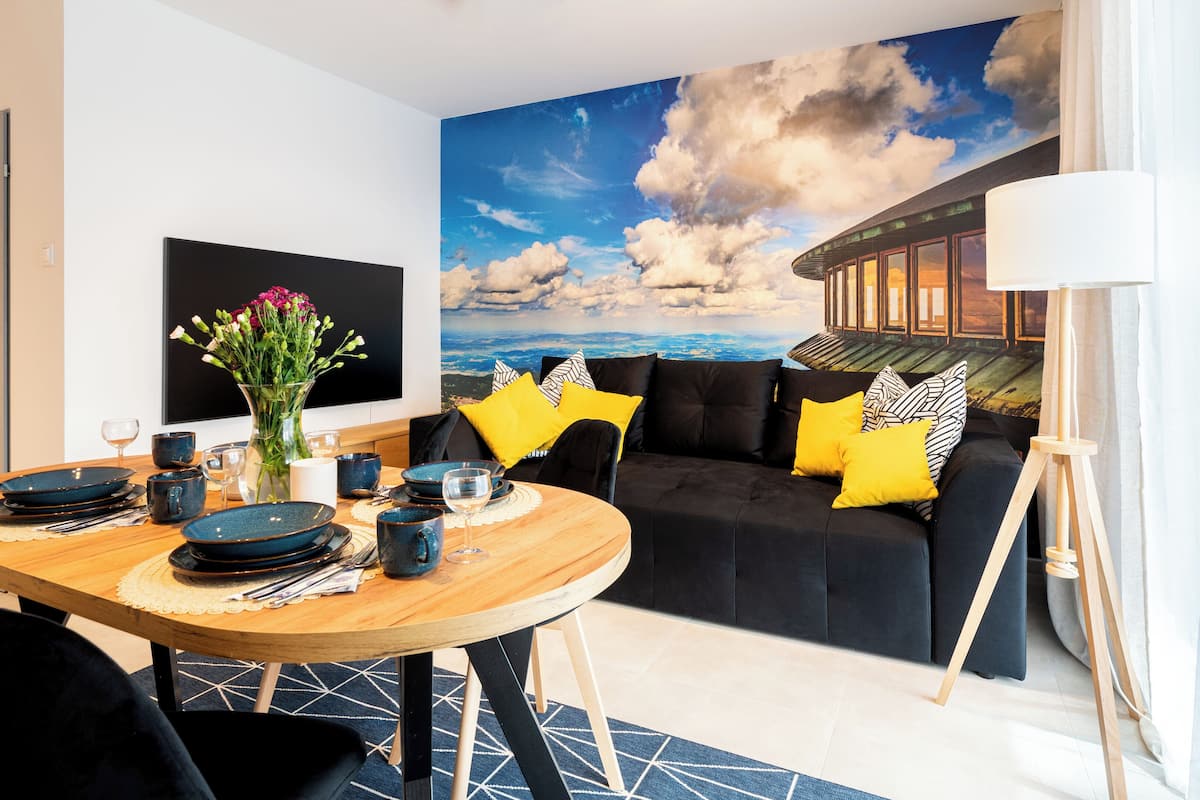 Een moderne woonkamer met een zwarte bank, kussens met een geel-zwart patroon, een houten eettafel met blauwe borden en glazen, en een grote muurschildering van een lucht en wolken.