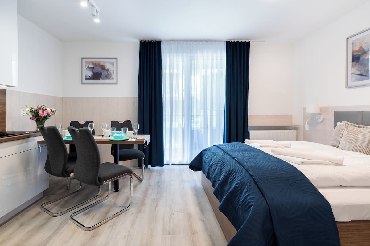 Een moderne hotelkamer met een bed, een bureau met stoelen, een kitchenette en een groot raam met gordijnen.