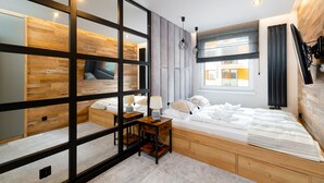 Een moderne slaapkamer met een houten hoofdbord, een groot bed, een nachtkastje met een lamp, en een aan de muur bevestigde televisie.