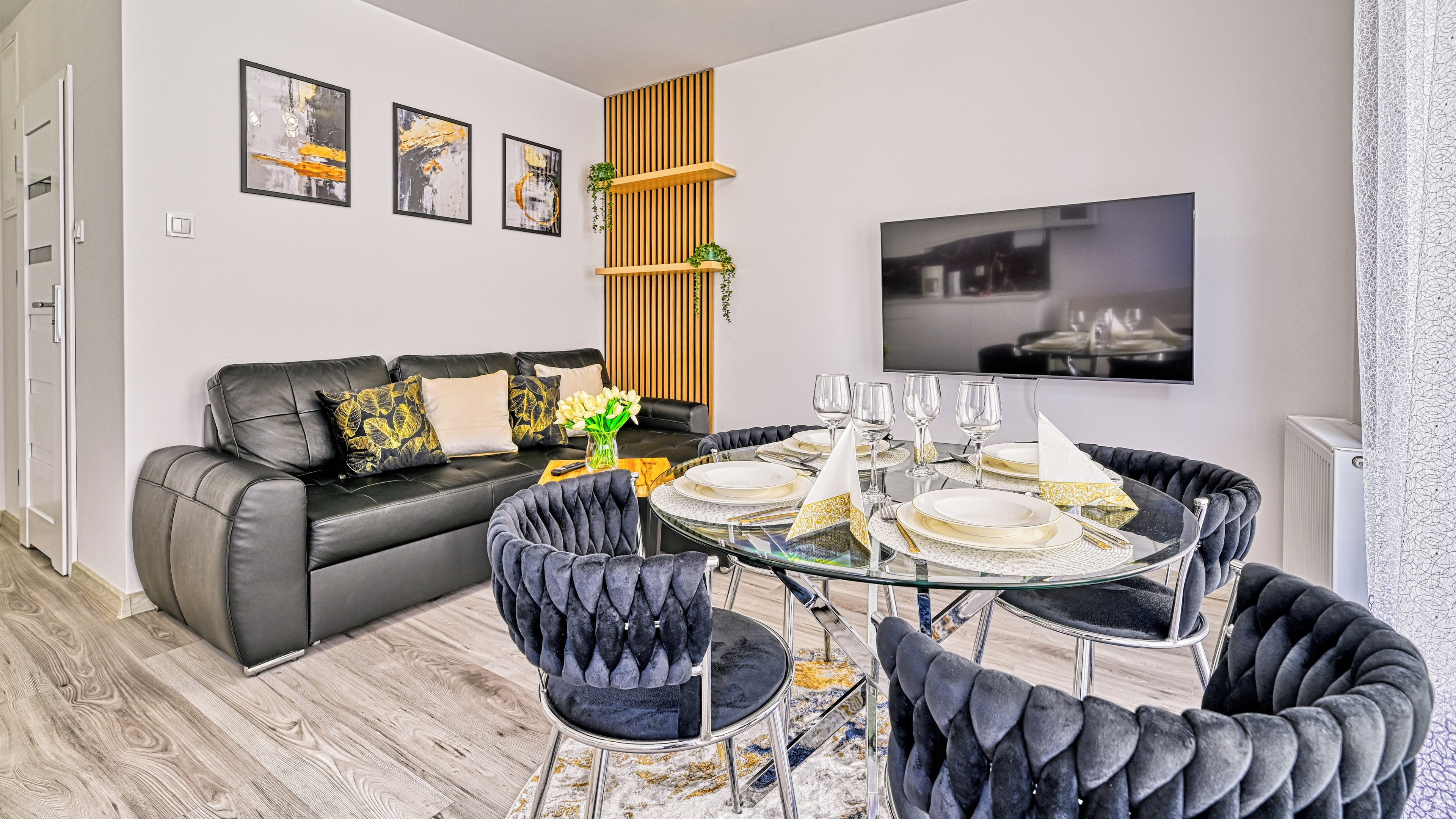 Una sala de estar moderna con un sofá de cuero negro, una mesa de comedor de vidrio preparada para cuatro personas y un televisor montado en la pared.