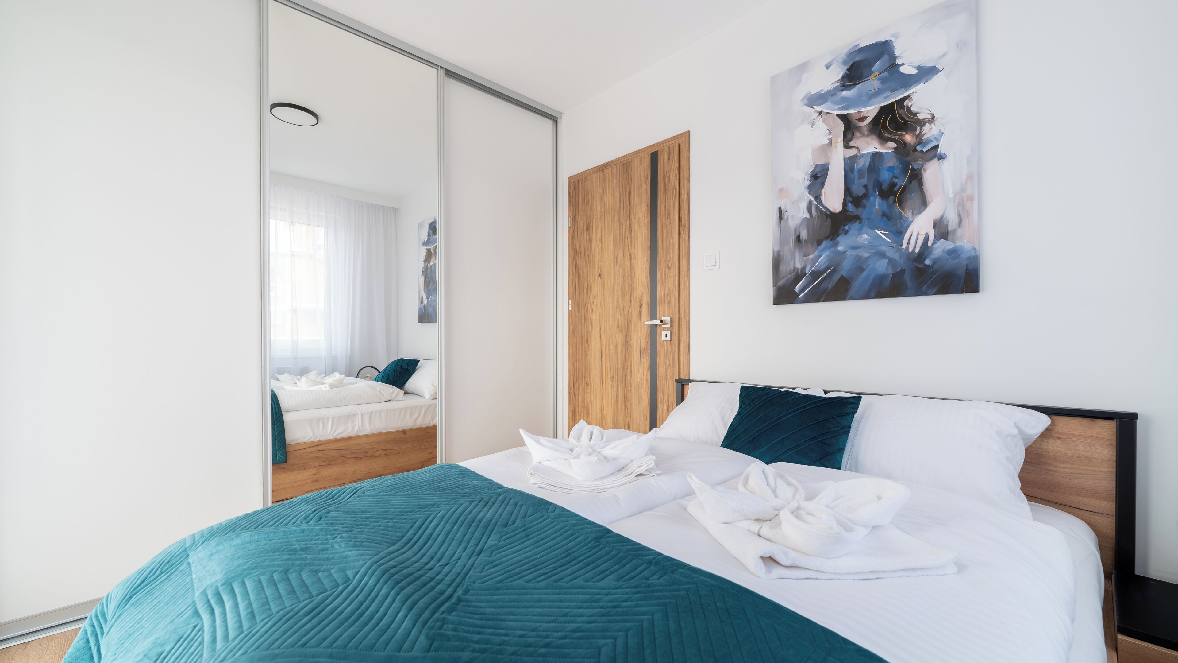 A modern hotel room with a large bed, a wooden wardrobe, a painting on the wall, and a sliding glass door leading to a balcony.