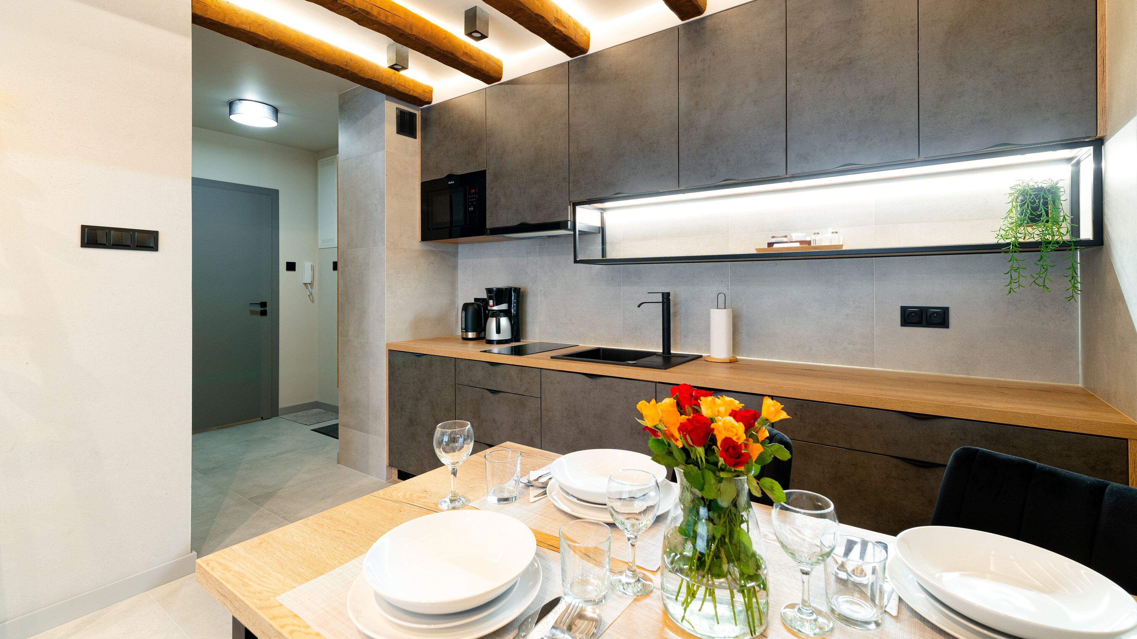 A modern kitchen with a built-in oven, a coffee maker, and a sink. There is a dining table set with plates, glasses, and a vase of flowers.