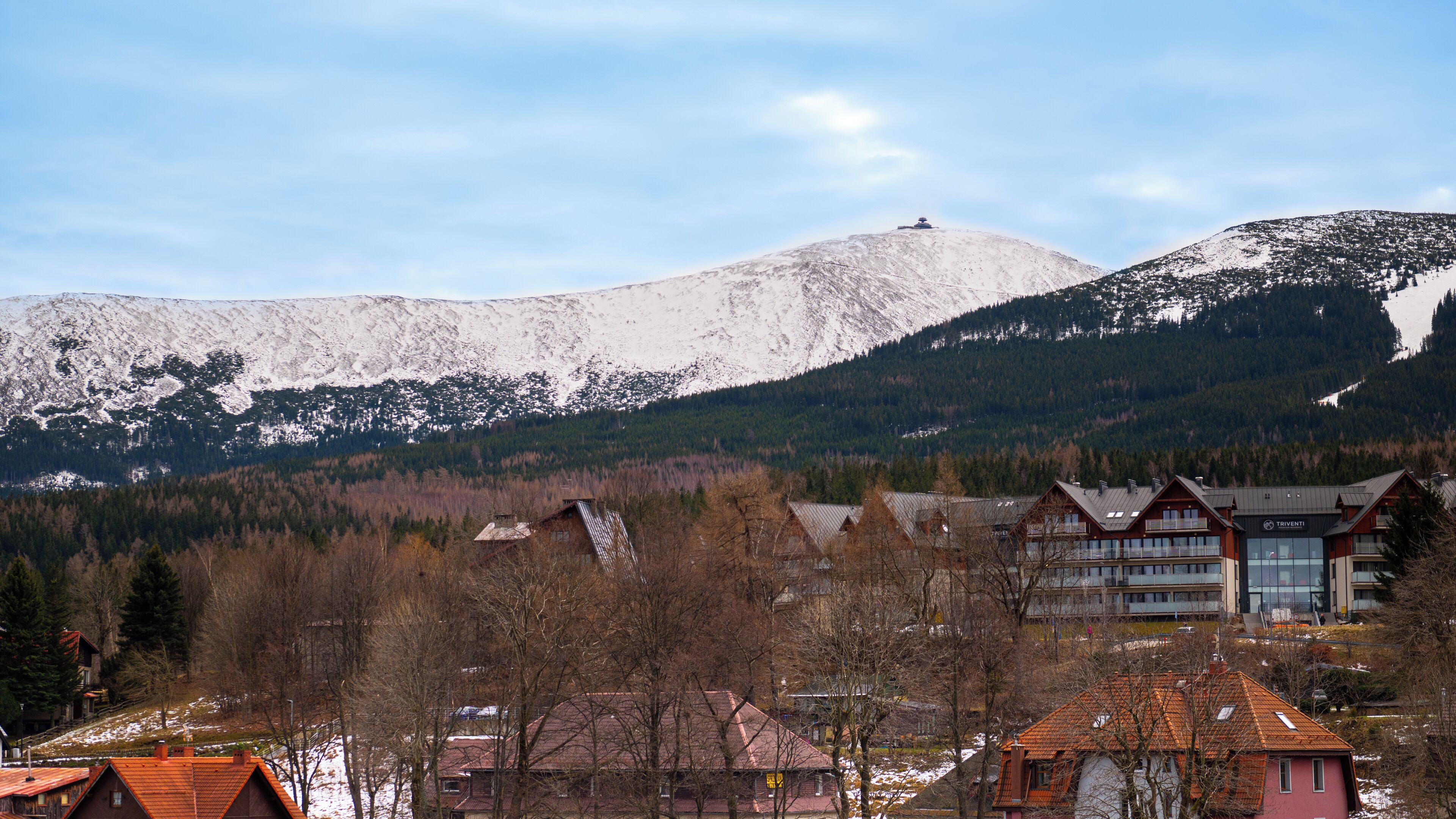Une montagne dont le sommet est coiffé de neige, une vallée boisée et un ensemble de bâtiments à l’architecture moderne.
