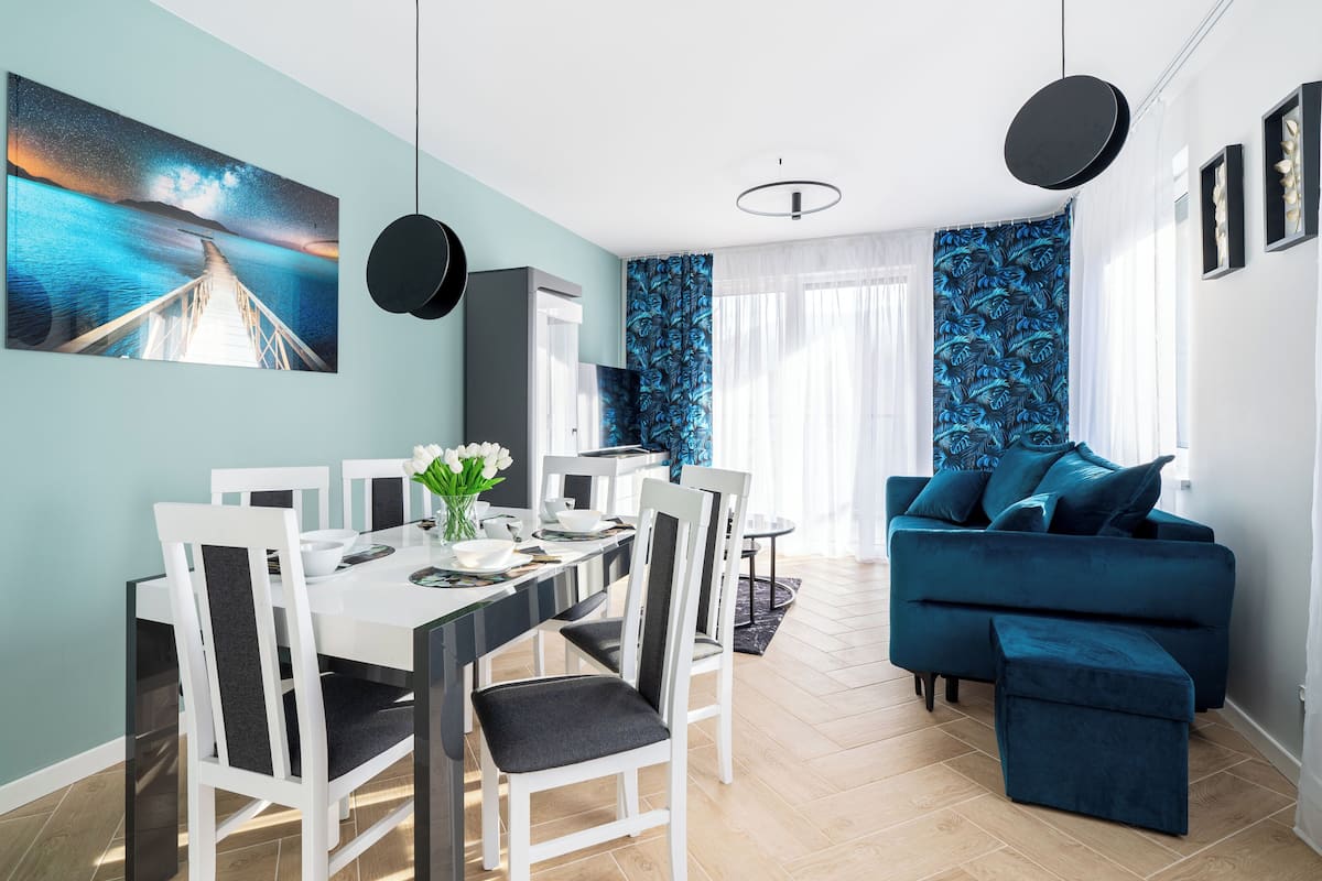 Een moderne woonkamer met een blauwe bank, een eethoek met een witte tafel en stoelen, en een groot schilderij van een nachthemel.