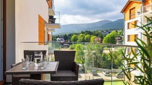 Een balkon met rieten meubels en een glazen tafel, met uitzicht op een woongebied met huizen en bergen op de achtergrond.