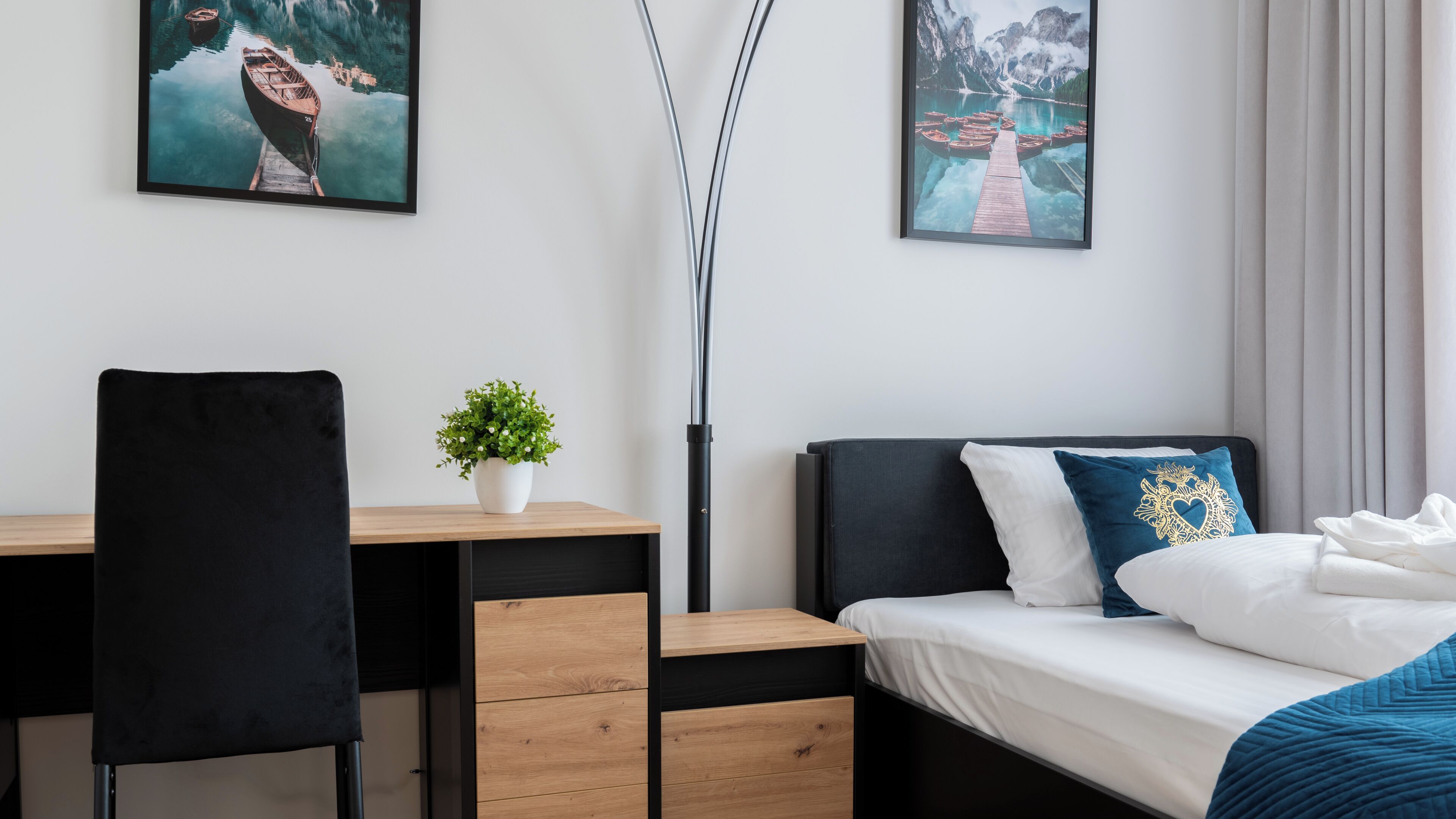 A modern hotel room with a wooden desk, a black chair, a bed with white and blue bedding, and two framed pictures on the wall.