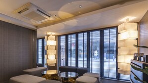 A modern hotel room with a seating area featuring a round table and four cushioned chairs, large windows with blinds, and a decorative light fixture.