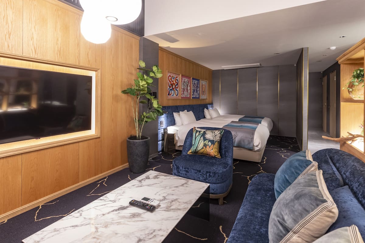 A modern hotel room with a large flat-screen TV, a marble coffee table, a sofa, a bed with blue bedding, a potted plant, and artwork on the wall.