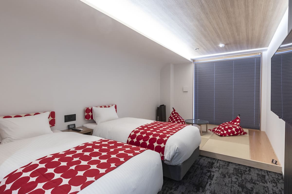 A modern hotel room with two beds, a small table, and a large window with blinds.