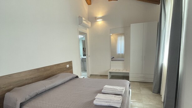 Une chambre avec un plafond en bois, un lit recouvert d’une couette grise, une table de chevet, une armoire et une salle de bain visible au fond.