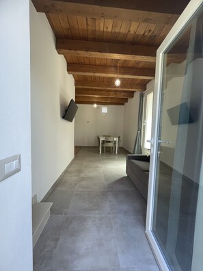 Une pièce avec un plafond en bois, une porte-fenêtre coulissante, un canapé gris, une petite table avec des chaises et un téléviseur fixé au mur.