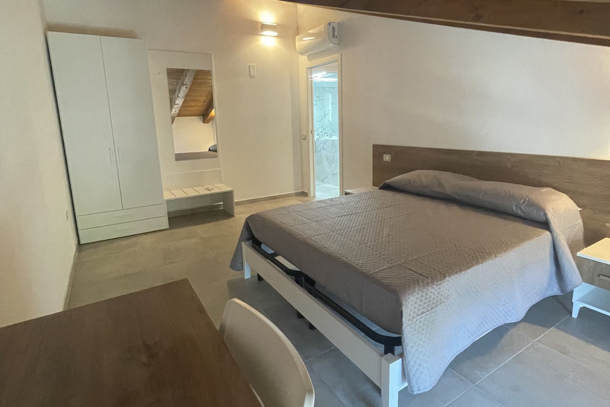 A bedroom with a bed, a desk, and a chair. There is a wardrobe and a door leading to another room.