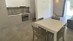 Une cuisine moderne avec des armoires blanches, un évier en acier inoxydable et un four encastré. On y trouve un coin repas avec une table et des chaises blanches. Le plafond est en bois avec des poutres apparentes.