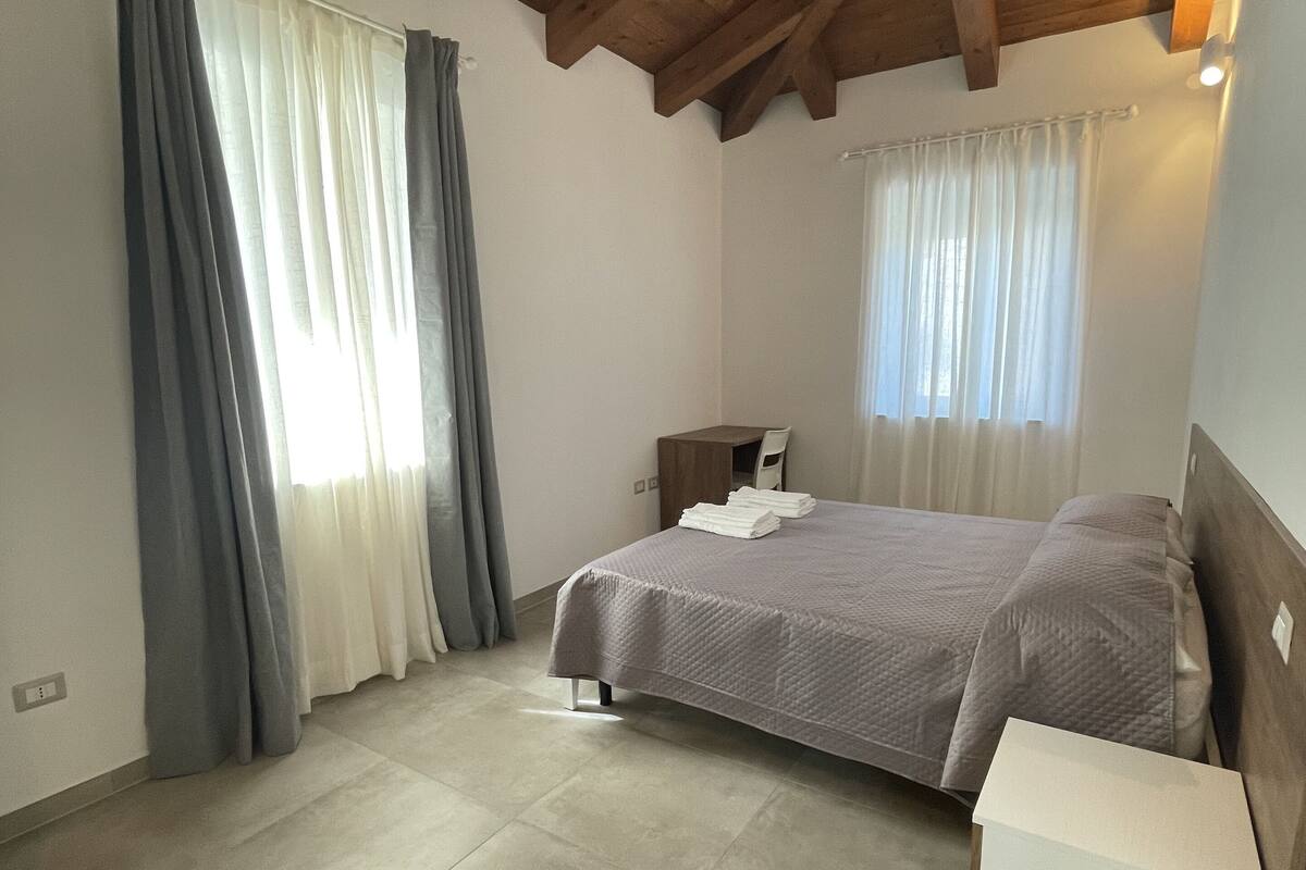 A bedroom with a bed, a nightstand, a bench, and a window with curtains.