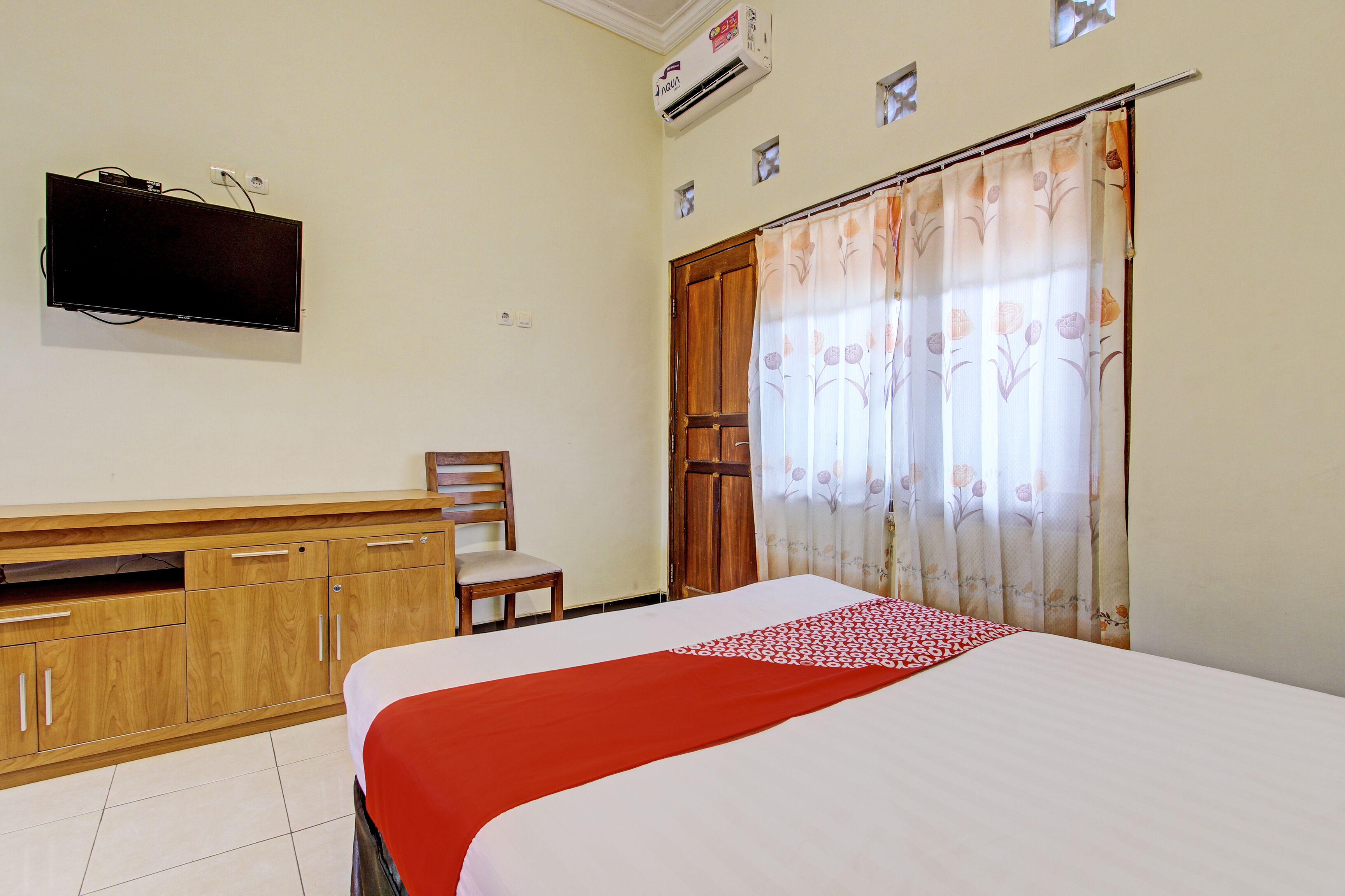 A hotel room with a bed, a TV mounted on the wall, a wooden wardrobe, a chair, and a window with curtains.