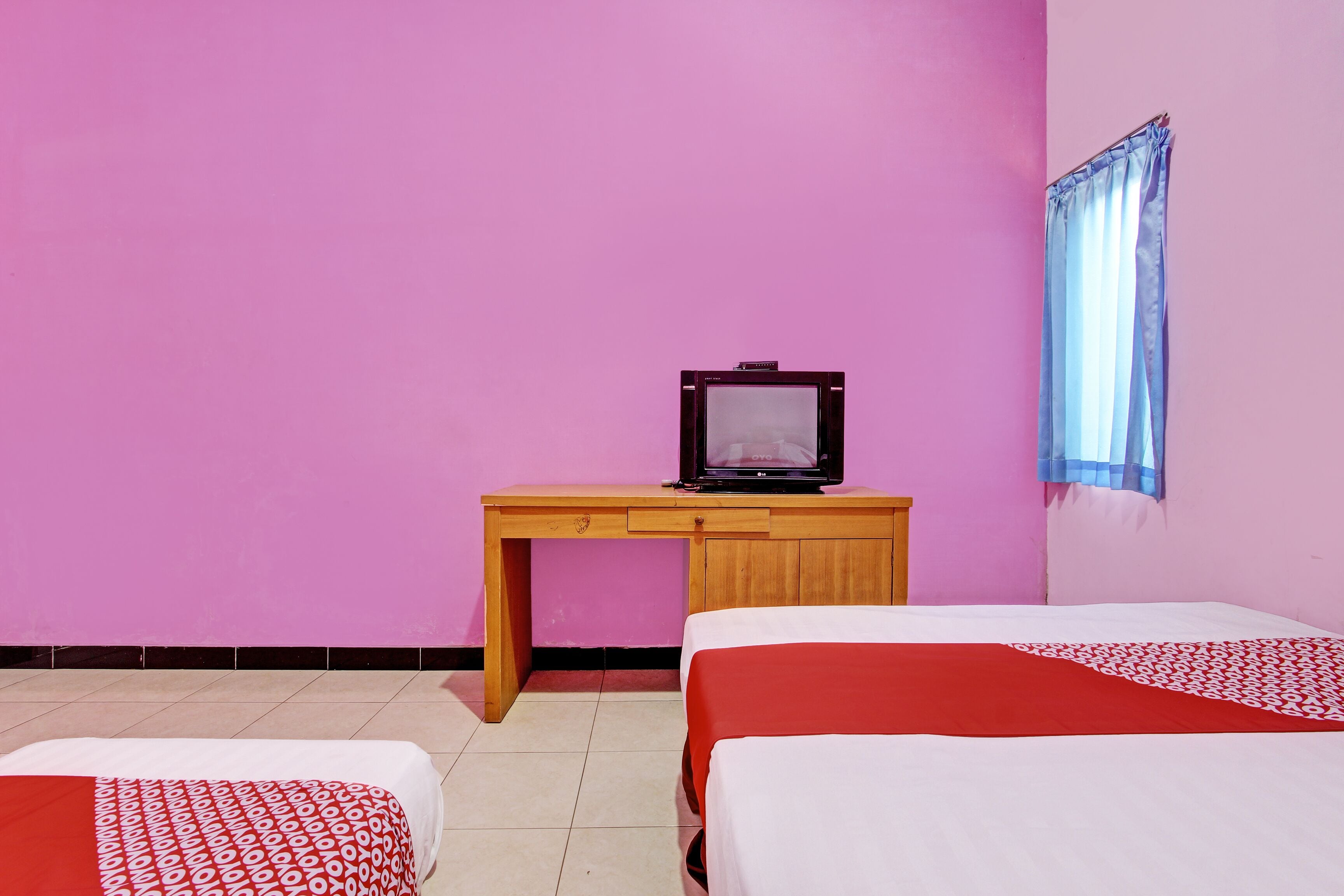 A room with two beds, a desk with a television, and a window with a curtain.