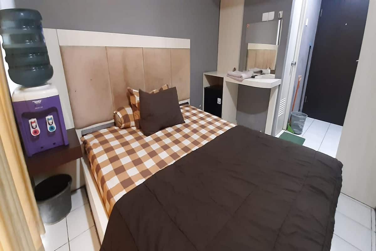 Ein Hotelzimmer mit Bett, Nachttisch, Wasserspender, Schreibtisch mit Spiegel und Mülleimer.