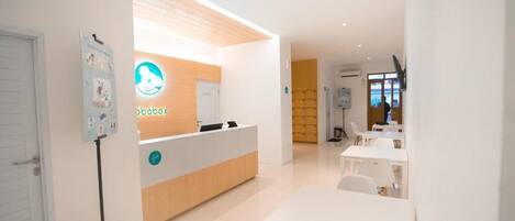 A modern reception area with a wooden reception desk, a white wall with a logo, and a seating area with tables and chairs.
