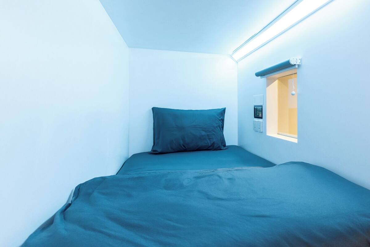 A small, single-bed room with a window, a wall-mounted thermostat, and a ceiling light.