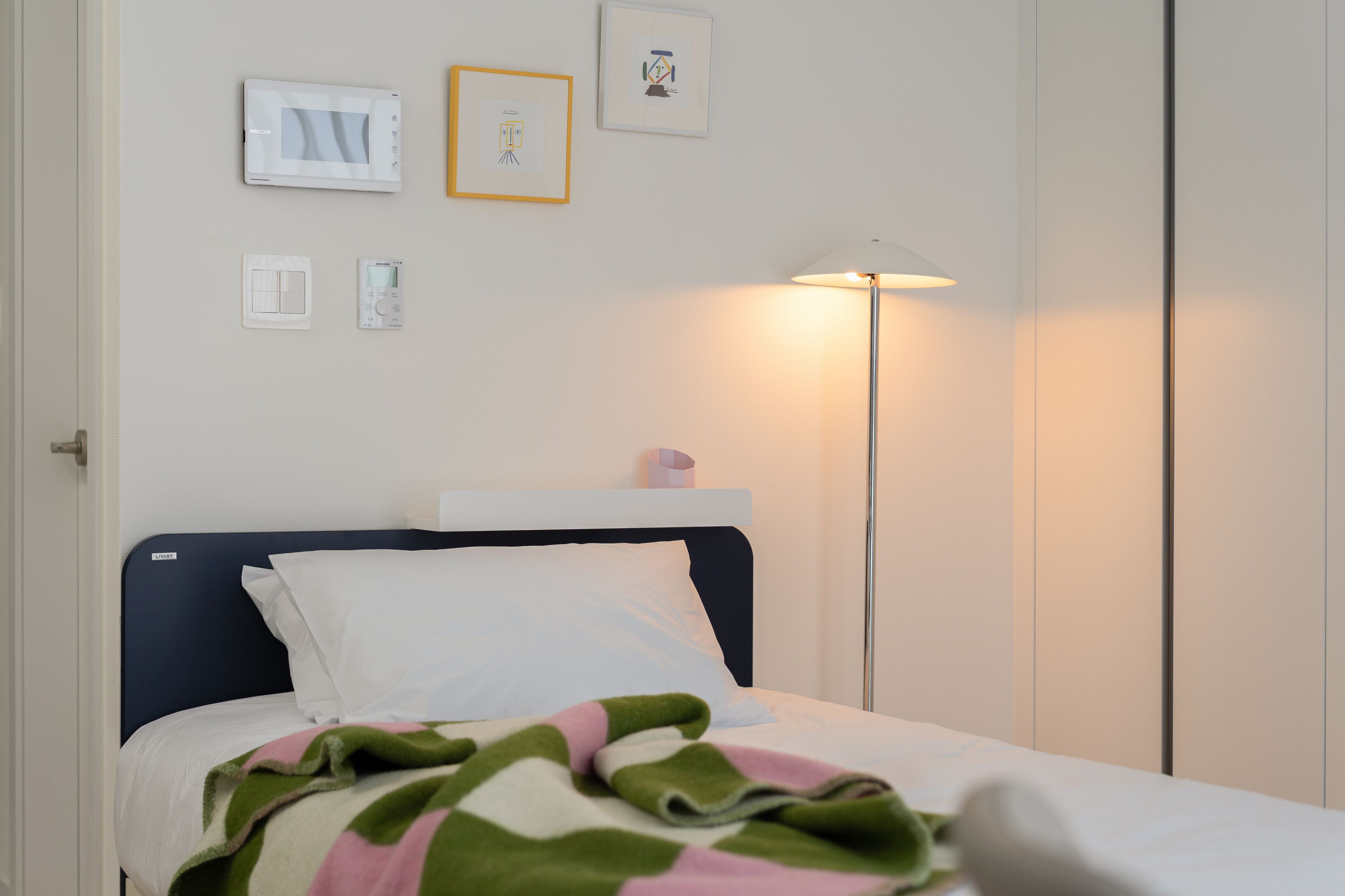 A neatly made bed with a striped blanket, a bedside lamp, and a wall-mounted thermostat.