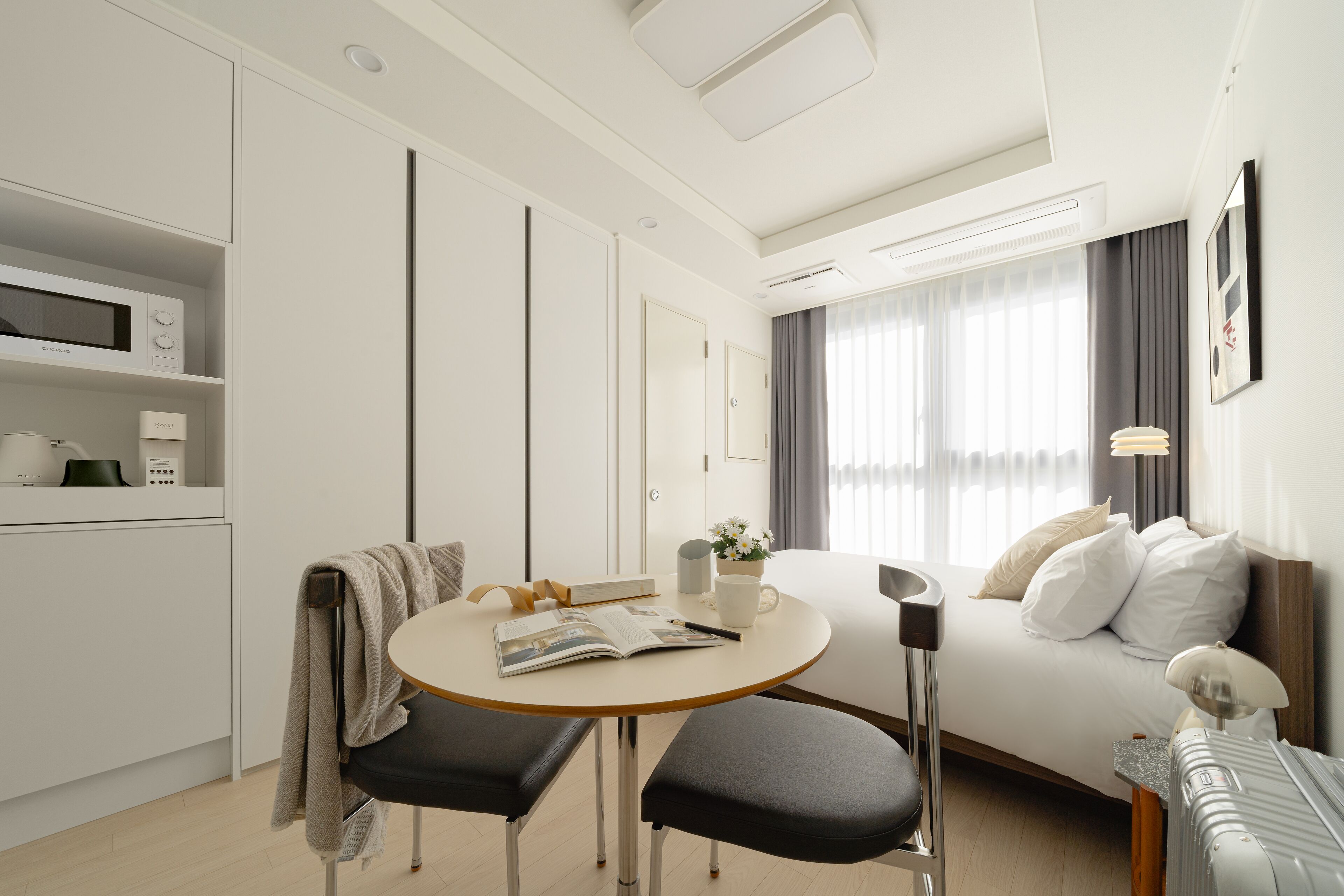 A modern hotel room with a bed, a round table with two chairs, a small kitchenette, and a large window with curtains.