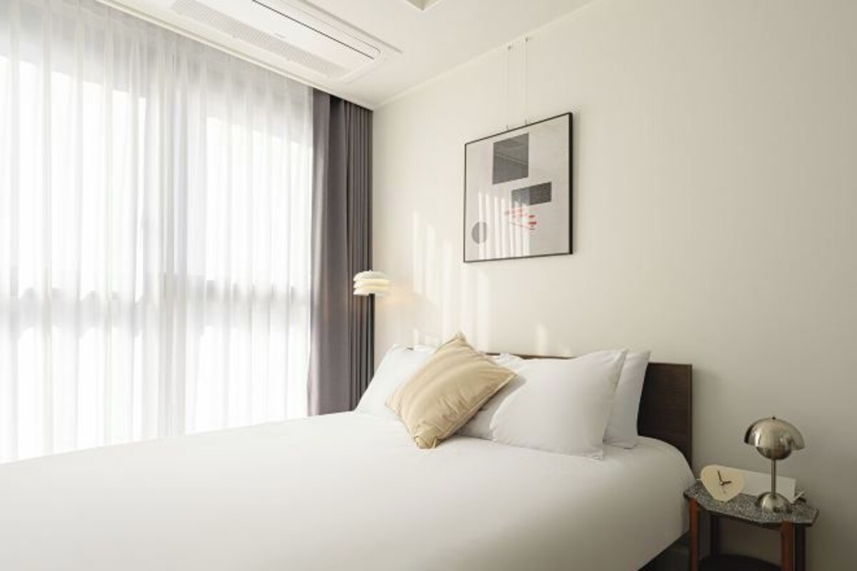 A hotel room with a bed, a bedside table with a lamp, a framed picture on the wall, and a window with curtains.