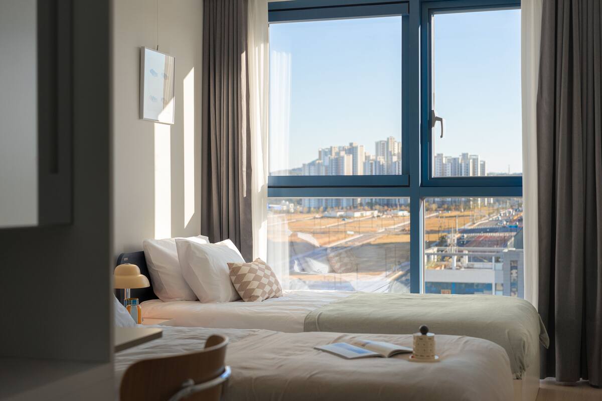 Una habitación de hotel con un ventanal grande, una cama, una mesita de noche y una silla.