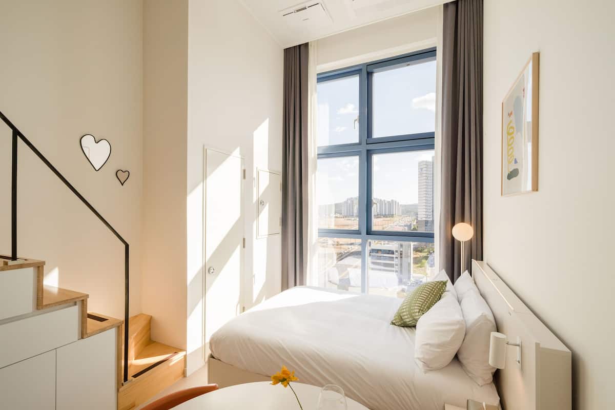 Una habitación de hotel moderna con un ventanal grande, una cama con ropa de cama blanca, una mesita con un florero y una escalera con escalones de madera.