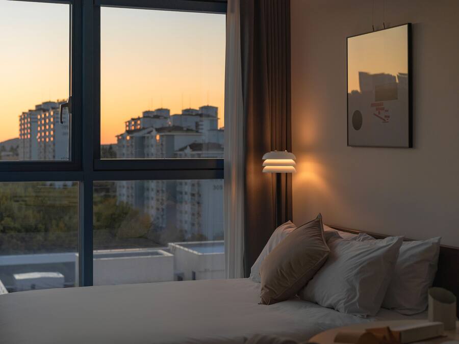 A bedroom with a large window, a bed with pillows, a bedside table with a lamp, and a framed picture on the wall.